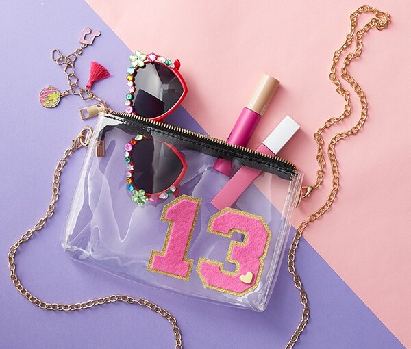 clear bag with chain with sunglasses and lip-gloss in it on a purple and pink background